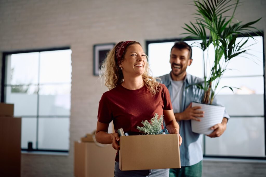 Smiling couple carrying moving boxes and a houseplant while moving into a new home.
