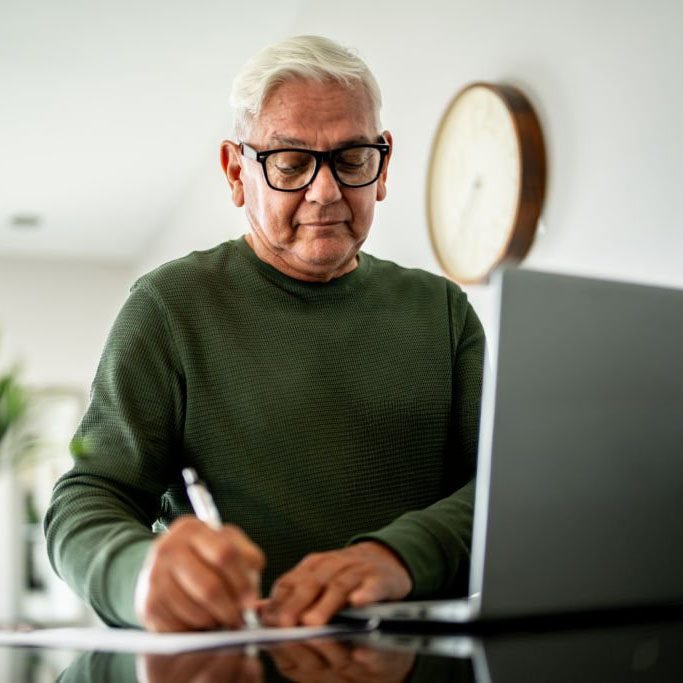 Homeowner reviewing documents and taking notes while working on a laptop at home.